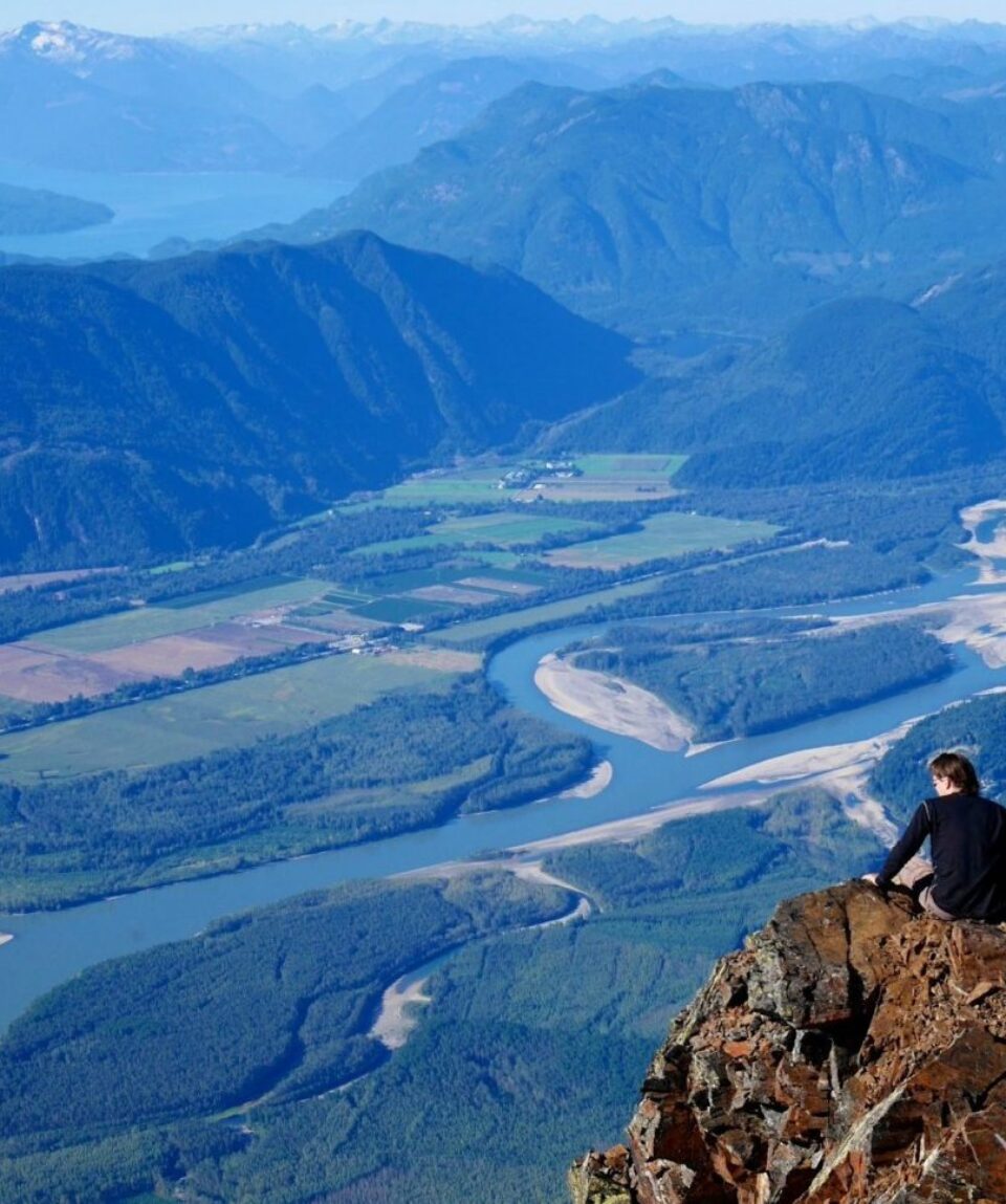 Man Sitting on Mountain Top with View of Valley, River, and Mountains. View of Fraser Valley and Harrison Hot Springs from Mount